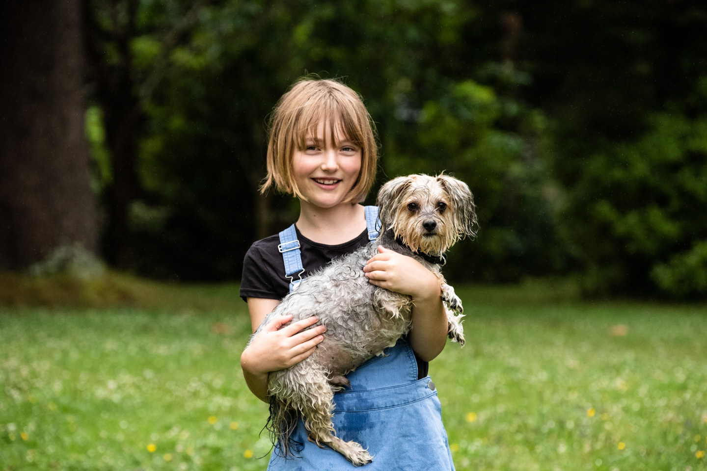 A young girl in dungarees holds her small scruffy dog in a park, both smiling at the camera with trees soft behind