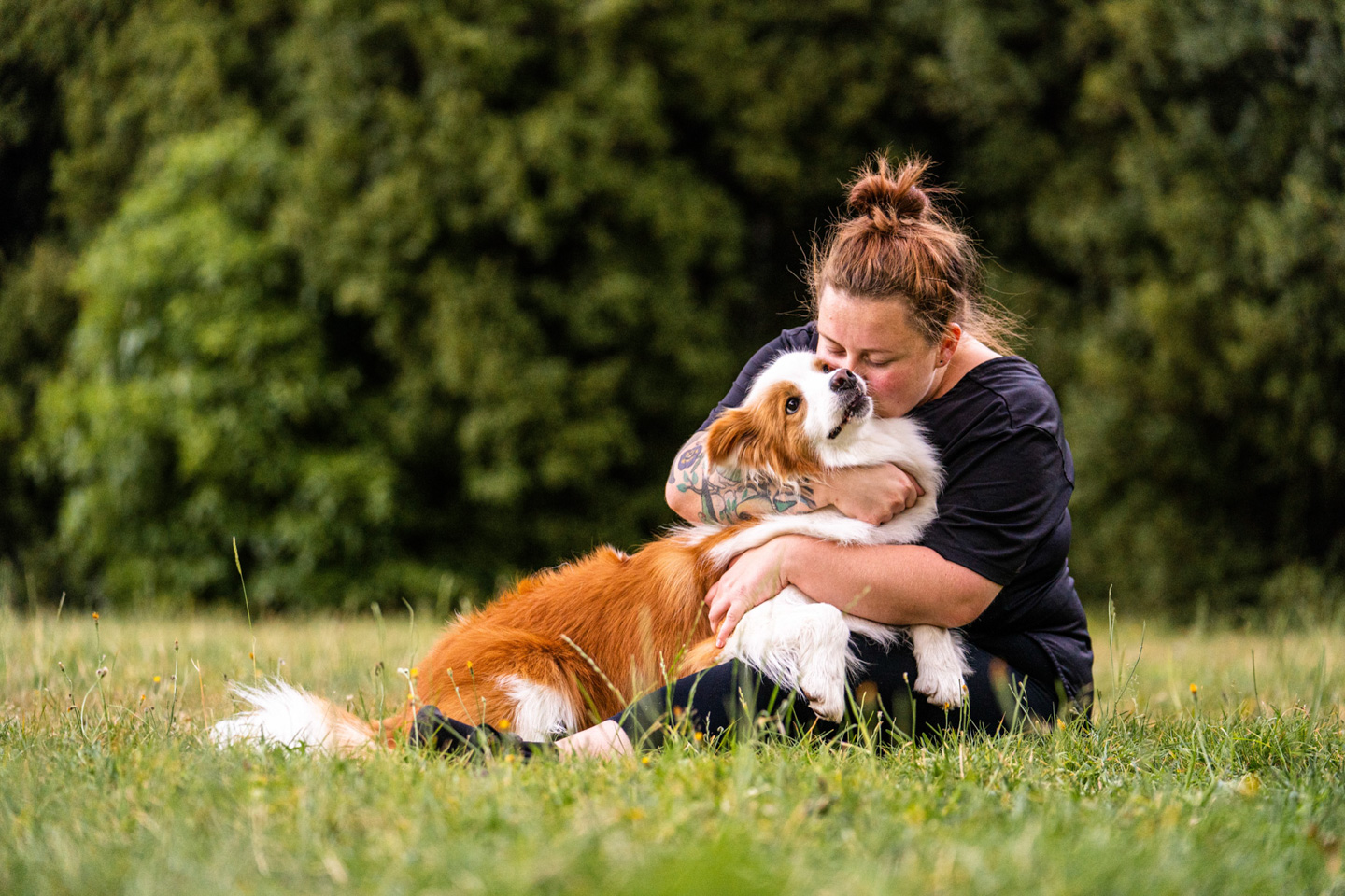 A woman sits on the grass cuddling her red and white dog, faces close together in a tender moment